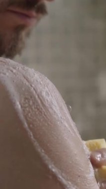 Dikey. Genç beyaz adamın duş alırken omuzlarından boynuna kadar sabunlu süngerle kendini temizlediği yakın plan. Yetişkin uzun saçlı erkek günlük temizlik ve vücut bakımını banyoda tek başına yapıyor. 