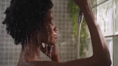 Yetişkin afro kadın saçlarını ve başını banyoda duş başlığıyla ıslatıyor. Genç Afro-Amerikalı güzel bir kadın evinde günlük temizlik yapıyor.