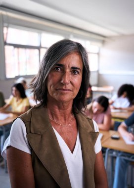 Dikey. Ciddi bir portre. Yetişkin bir beyaz kadın öğretmen. Sınıfta kendine güvenerek poz veriyor. Orta yaşlı bir öğretmen, kameraya ciddi bakıyor. Sınıfta dikkati dağınık öğrenciler var.