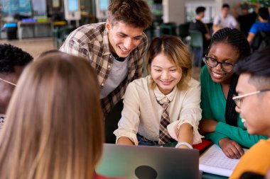 Akademi çalışma odasında oturmuş dizüstü bilgisayara gülümseyen çok ırklı bir grup genç öğrenci. Çeşitli üniversite öğrencileri dizüstü bilgisayarlarını kullanarak okul projeleri üzerinde çalışıyorlar.
