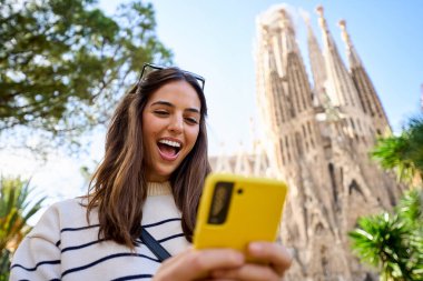 Barcelona 'da Sagrada Familia' nın önünde cep telefonu kullanan genç bir bayan arkadaş. Avrupa 'daki güzel beyaz kızlar tatilin tadını çıkarıyorlar ve gülümsüyorlar. Komik turizm insanları gülümser.