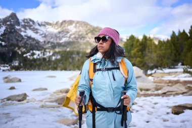 Sırt çantası ve yürüyüş direkleriyle donatılmış, karlı bir orman patikasının tadını çıkaran kadın yürüyüşçü, sakin kış manzarasını kucaklıyor.