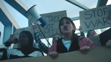 Megafonlu sarışın kadın çeşitli grup eylemcileriyle protesto ediyor. Dünya 'daki savaşlara karşı sloganlar atıyorlar. Elinde barışçıl pankartlar ve açık havada duran duman fişekleri var. 