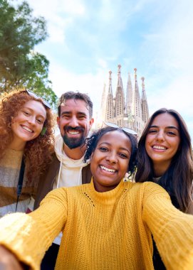 Vertical. Group diverse funny young tourist friends posing piggyback with hands joined in air taking selfie together in front of Sagrada Familia in Barcelona. Happy people enjoy holidays outdoor clipart