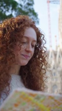 Dikey. Elinde harita olan genç kızıl kadınlar Barcelona 'da Sagrada Familia' nın önünde seyahat ediyorlar. Avrupa 'daki güzel beyaz kız tatilin tadını çıkarıyor ve gülümsüyor. Eğlence turizmi insanları güldürür.