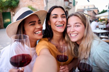Üç neşeli, çok kültürlü bayan arkadaş gülümseyip selfie çekerken Avrupa 'nın büyüleyici bir kasabasında bir restoranda kırmızı şarap içiyorlar.