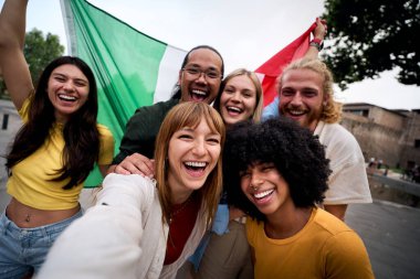 Bir İtalyan bayrağı tutarken selfie çeken, gülen, kutlama yapan ve şehir merkezinde birlikte eğlenen çok ırklı, neşeli genç bir grup.