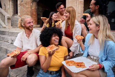 Bir grup neşeli, etnik çeşitliliğe sahip genç arkadaş tarihi bir binanın önünde oturmuş birlikte sokak pizzası yiyip gülüyorlar.