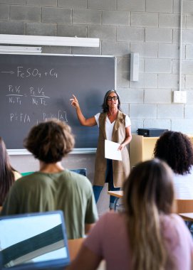 Dikey. Sınıfta not alan bir grup çok ırklı öğrencinin arkasından bakın. Öğretmen tahtada duruyor, bir ders veriyor, odaklanmayı ve bilgi alışverişini vurguluyor.