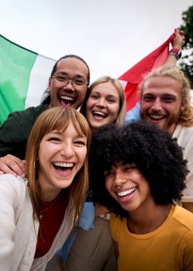 Dikey. Bir İtalyan bayrağı tutarken selfie çeken, gülen, kutlama yapan ve şehir merkezinde birlikte eğlenen çok ırklı, neşeli genç bir grup.