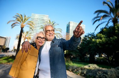 Mutlu emekli çift Barselona, İspanya 'da güneşli bir günün keyfini çıkarırken, adam karısını kucaklarken ilgi çekici bir şeye işaret ediyor, neşe ve keşif anını paylaşıyor.
