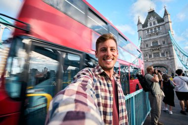 Londra 'da selfie çeken genç bir adam kırmızı çift katlı bir otobüsle oradan geçiyor ve arka planda kule köprüsü var. İngiltere' nin başkentine yaptığı yolculuğun tadını çıkarıyor.