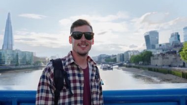 Genç adam neşeli bir ifadeyle kameraya bakıyor. Londra ufuk çizgisi arka planda görülebilir, sahneye kentsel ve modern bir atmosfer ekliyor.