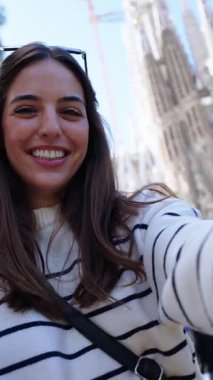 Dikey. Genç beyaz kadınlar Barcelona 'da Sagrada Familia' nın önünde selfie çekiyorlar. Avrupa şehrinde gülümseyen güzel kız tatilin tadını çıkarıyor ve gülümsüyor. Komik turizm insanları.