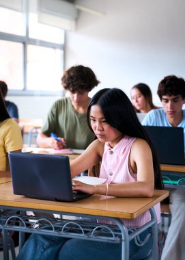 Dikey. Gülümseyen Asyalı liseli kız sınıfta dizüstü bilgisayar kullanıyor. Sınıfta bir grup mutlu sınıf arkadaşı. Eğitim ve teknoloji. Yüksek kalite fotoğraf