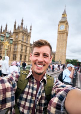 Dikey. Sırt çantalı genç beyaz adam Westminster Köprüsü, Londra 'da akıllı telefonuyla selfie çekiyor. Ben ve parlamento binaları arka planda, yaz tatillerinin tadını çıkarıyor.