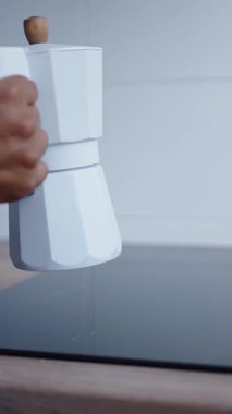 Dikey video. Siyah indüksiyon sobasının üzerine ahşap saplı beyaz moka tenceresi koyan kadın, zarif, modern mutfak alanında taze kahve demliyor.