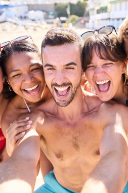 Mayolu neşeli turistlerden oluşan dikey bir grup, yolculuk hatıraları için sahilde mutlu bir selfie çekiyor. Çeşitli arkadaşlar gülüyor, boş zamanların tadını çıkarıyor ve yaz tatillerinde eğleniyorlar.