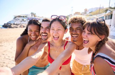 Sahildeki çok ırklı genç insanlar sosyal ağlarda paylaşmak için mayo giyip selfie çekiyorlar. Arkadaşlar gülümseyerek yaz tatilinin tadını çıkarıyor.