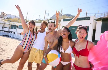 Mayo giymiş bir grup genç insan kumsalda fotoğraf çektirmek için kollarıyla poz vererek gülüyor, güneşli gökyüzünün altında boş zamanların ve yaz yolculuğunun tadını çıkarıyorlar. Nesiller z arkadaş ilişkileri