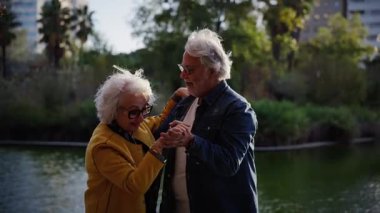 Neşeli emekli çift, canlı bir şehir parkında sakin bir gölün kenarında romantik bir dansın keyfini çıkarırken, ebedi aşklarını ve aktif yaşam tarzlarını kutluyorlar.
