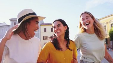 Güneşli bir yaz gününde hareketli bir kasaba meydanında el ele tutuşup keyif ve dostluk saçan üç neşeli genç kadın turist.