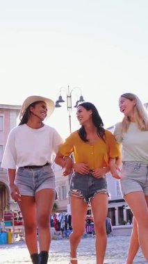 Dikey. Güneşli bir yaz gününde hareketli bir kasaba meydanında el ele tutuşup keyif ve dostluk saçan üç neşeli genç kadın turist.