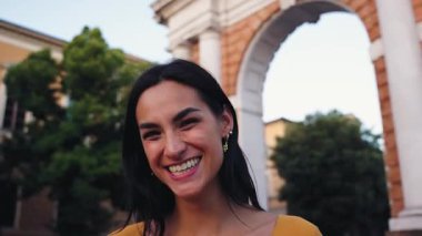 Uzun siyah saçlı, altın küpeli, turuncu gömlekli ve kentsel bir kemer yolunun önünde dikilirken gülümseyen neşeli Latin bir kadının portresi.