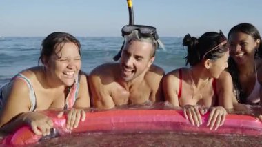 Bir grup kahkaha ve çeşitli neşeli genç arkadaşlar deniz kıyısında şişme yatakta fotoğraf için poz veriyorlar. Çok ırklı, heyecanlı genel dostlar güneşli tatillerin ve kumsalda boş vakitlerin tadını çıkarıyorlar.