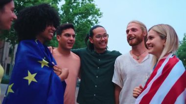 ABD bayrağı ve Avrupa Birliği bayrağını tutan neşeli bir grup genç el sıkışıyor. Görüntü, kültürler arasındaki dostluğu, birliği ve işbirliğini simgeliyor.