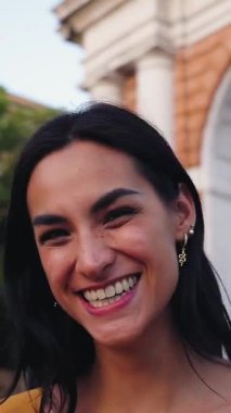 Dikey. Uzun siyah saçlı, altın küpeli, turuncu gömlekli ve kentsel bir kemer yolunun önünde dikilirken gülümseyen neşeli Latin bir kadının portresi.