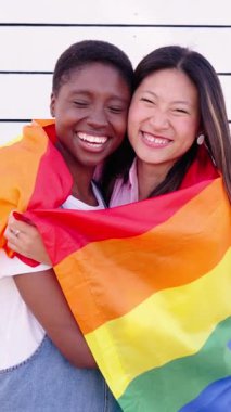 Dikey. Genç lezbiyen çift, eşcinsel gurur bayrağıyla sarılıyor. Mutlu arkadaşlar gülümseyen kameraya bakıp beyaz arka plana sarılıyorlar. Asya ve Afrika birleşik homoseksüel aşkı
