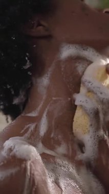 Dikey. Genç Afrikalı Afrikalı Amerikalı kadın banyo süngeriyle vücuduna duş jeli sürüyor, kendini rahatlama hissine sokuyor. Kişisel bakım anı onun evdeki doğal güzelliğini vurguluyor.