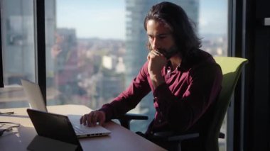 Modern ofis masasında dizüstü bilgisayarla çalışan uzun saçlı profesyonel Latin iş adamı. Etrafı parlak şehir manzarası ve geniş pencerelerle çevrili. Dijital görevlere odaklanıyor.
