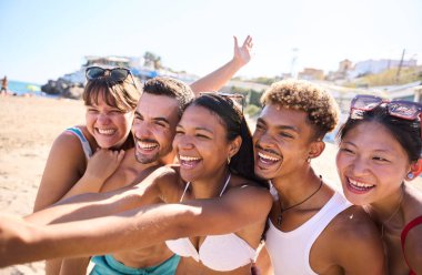 Mayo giyen bir grup Z nesli kumsalda mutlu mesut selfie çekiyorlar, neşeli ve büyük gülücükler saçıyorlar. Çeşitli gençler yaz tatili maceralarının tadını çıkarıyorlar.