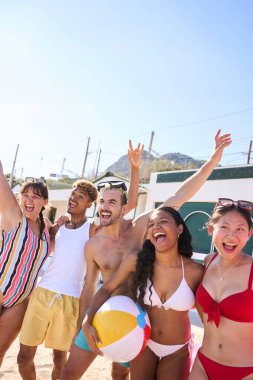Dikey. Mayo giymiş bir grup genç insan kumsalda fotoğraf çektirmek için kollarıyla poz vererek gülüyor, güneşli gökyüzünün altında boş zamanların ve yaz yolculuğunun tadını çıkarıyorlar. Gen z arkadaş ilişkisi