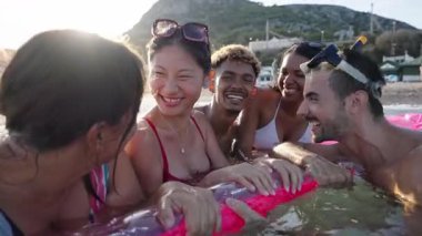 Bir grup çok ırklı insanın bikinisiyle gülüp eğlendiği ve deniz kıyısındaki şişme bir paspasın üzerinde dalış gözlüğü taktığı bir yaz havası. Z nesli, boş zaman ve tatil zamanı.