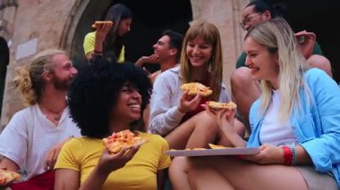 Bir grup neşeli, çok ırklı genç arkadaş yaz tatilleri boyunca antik taş basamaklarda oturup pizza dilimlerinin tadını çıkarıyorlar.
