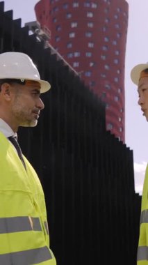 Dikey. Açık havada iki çok kültürlü mimarla toplantı takım liderleri projeyi tokalaşarak bitiriyorlar. Çeşitli resmi profesyonel mühendisler, arka planda binalar bulunan güvenlik giysileri içinde selamlıyorlar.