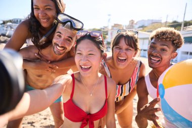 Mayo giymiş neşeli bir grup genç arkadaş sahilde komik bir selfie çekiyorlar, sırtüstü uzanıyorlar, güneşli gökyüzünün altında gülümsüyorlar. Nesiller boyunca insanlar yaz tatilinde eğleniyorlar.