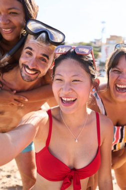 Dikey. Mayo giymiş neşeli bir grup genç arkadaş sahilde komik bir selfie çekiyorlar, sırtüstü uzanıyorlar, güneşli gökyüzünün altında gülümsüyorlar. Nesiller boyunca insanlar yaz tatilinde eğleniyorlar.