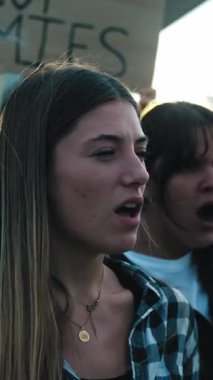 Dikey. Çeşitli gruptan genç sarışın kadın yumrukları havada savaşa karşı öfkeli bağırışları protesto ediyor. Dünya Barış Yürüyüşü gösterilerinde savaş karşıtı pankartlarla yürüyen farklı aktivistler