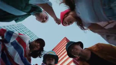 Çeşitli kadın ve erkekler bir çember oluşturup kameraya bakıyor ve dans ediyorlar. Arka planda modern bir bina var.