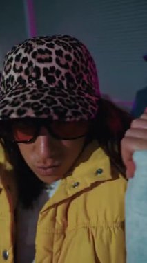 Dikey. Kendine güvenen genç bir kadın neon ışıklı şehir ortamında hip hop dansı yapıyor. Ekibi renkli arka planda ilerlerken ön planda duruyor.