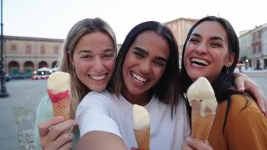 Üç neşeli bayan arkadaş bir yaz gününün tadını çıkarıyorlar, gülerken ve dondurma yerken bir yandan da şehir meydanında selfie çekiyorlar.
