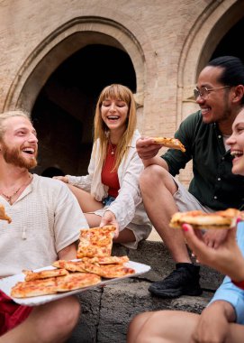 Dikey. Neşeli genç arkadaşlar birlikte lezzetli pizzaların tadını çıkarıyorlar, gülüyorlar ve neşeli anları paylaşıyorlar şehir hayatı dolu bir şehir ortamında taş basamaklarda otururken.