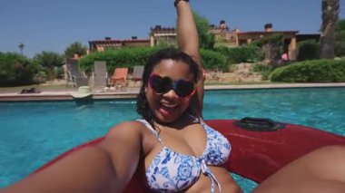 Afro-Latin bir kadın havuzda kendini videoya çeken bir selfie çekiyor, sonra iki arkadaş şişme bir yaz gecesinde ona katılarak gülüyor, güneşin altında neşeli bir yaz anını paylaşıyor. İnsanlar yaz havasını seviyor.