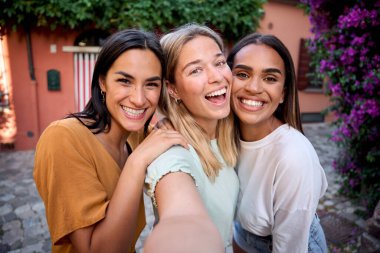 Üç neşeli, çok ırklı bayan arkadaş yaz tatillerini canlı çiçekler ve tarihi binalarla çevrili büyüleyici bir italyan kasabasında geçirirken selfie çekiyorlar.