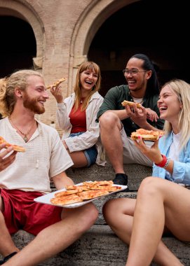 Dikey. Neşeli, çoklu etnik kökenli genç arkadaşlar gülüyor ve lezzetli pizzaları paylaşıyorlar. İtalya 'da çarpıcı bir tarihi binanın önünde oturmuş birlikte kaygısız bir anın tadını çıkarıyorlardı.