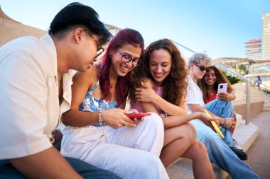 Bir grup neşeli genç öğrenci bir şehirde basamaklarda oturuyor. Akıllı telefondan gülüp komik içerikleri paylaşıyorlar. Birlikte geçirdikleri zamanın tadını çıkarıyorlar ve teknolojiyi eğlence ve bağlantı için kullanıyorlar.